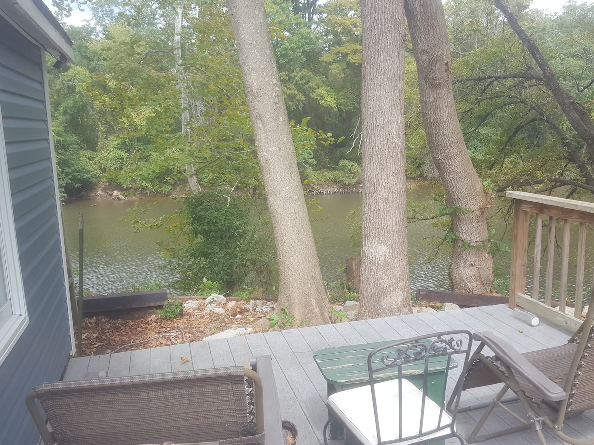A private deck overlooks a serene river, surrounded by tall trees. Outdoor seating includes a table and chairs, creating a relaxed space for enjoying nature. The wooden deck is bordered by gravel and natural foliage, offering a tranquil atmosphere.