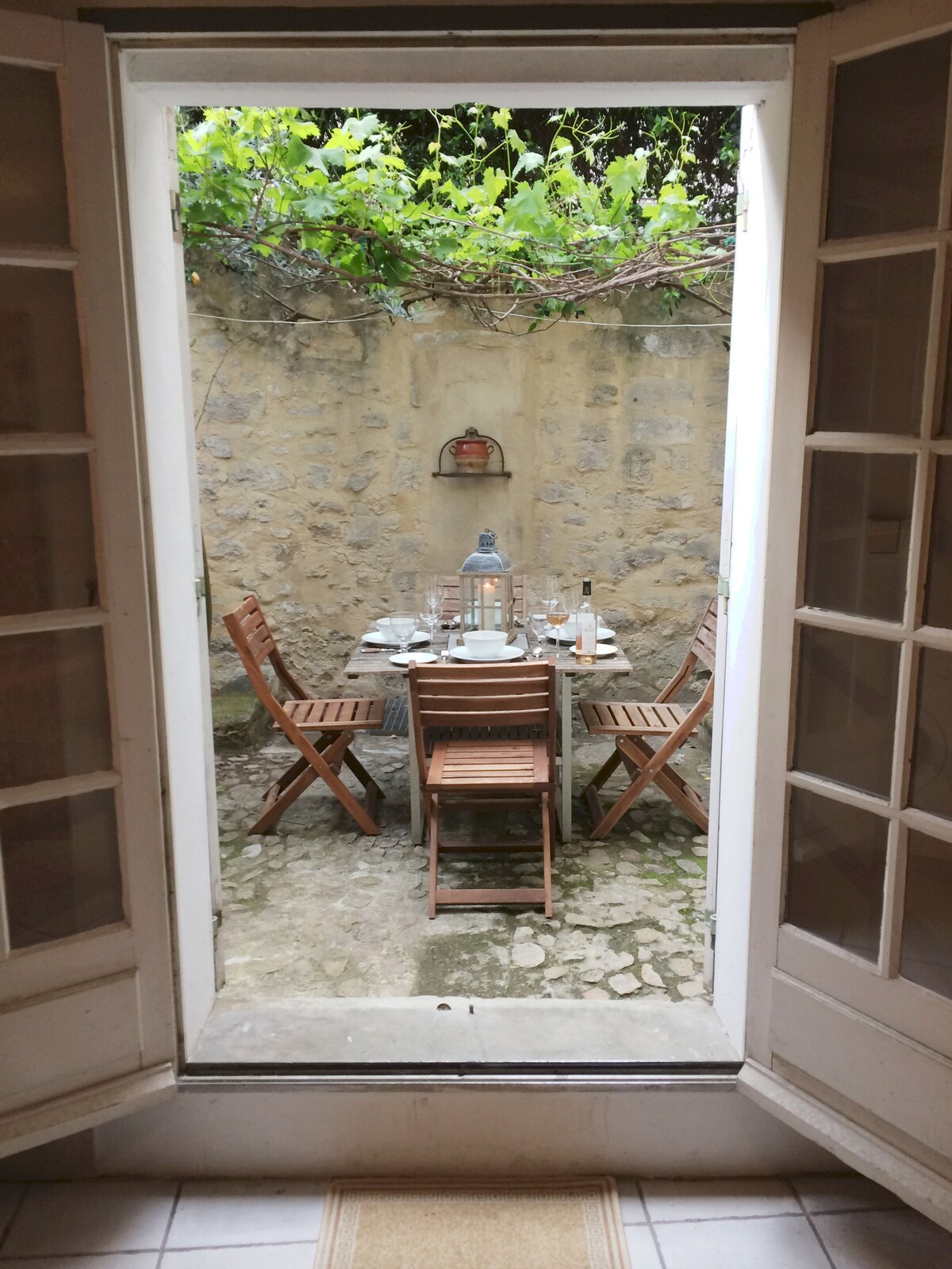 A charming cobbled courtyard is framed by double doors, featuring a wooden dining table set for a meal. Surrounding the table are several wooden chairs, with lush greenery visible above the stone walls, creating a tranquil al fresco dining space.