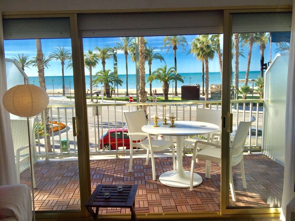 A private terrace features a round table surrounded by four white chairs, overlooking a palm-lined promenade and the beach. Large glass doors provide direct access to the outdoor space, allowing natural light to fill the interior.