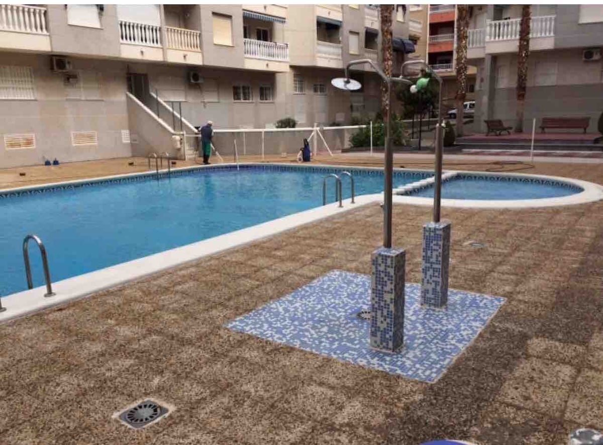 A sizable pool area is surrounded by a textured tiled deck. Two outdoor showers are positioned near the pool, offering easy access from the water. Sun loungers are organized along one side, while the surrounding buildings provide a sheltered environment.