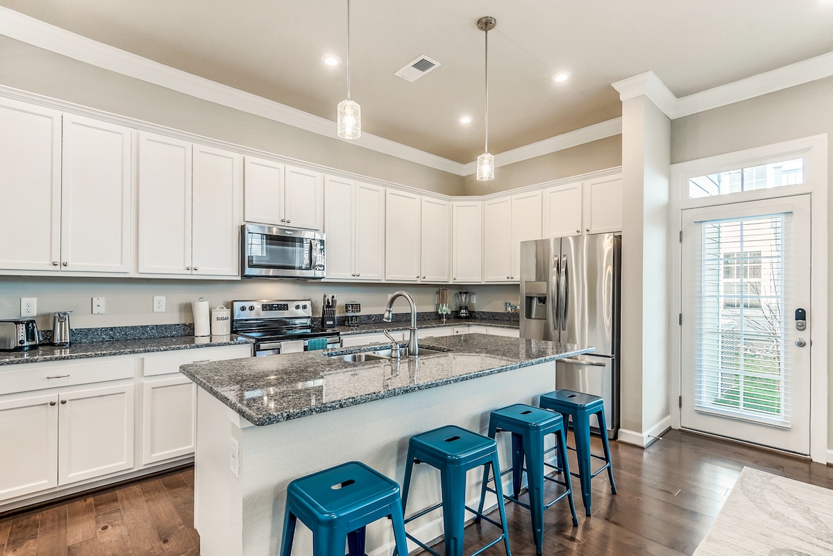 A spacious kitchen features a central island with a granite countertop and seating for four on blue stools. Stainless steel appliances, including a fridge and oven, are positioned along the white cabinetry. Natural light fills the room through glass doors leading to the outside.