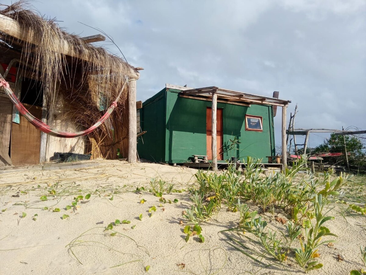 A rustic green cabin is situated among sandy terrain, with sparse vegetation and a hammock visible nearby. The structure features a red door and a small window, surrounded by a natural beach atmosphere. Distant elements of another building can be seen to the left.
