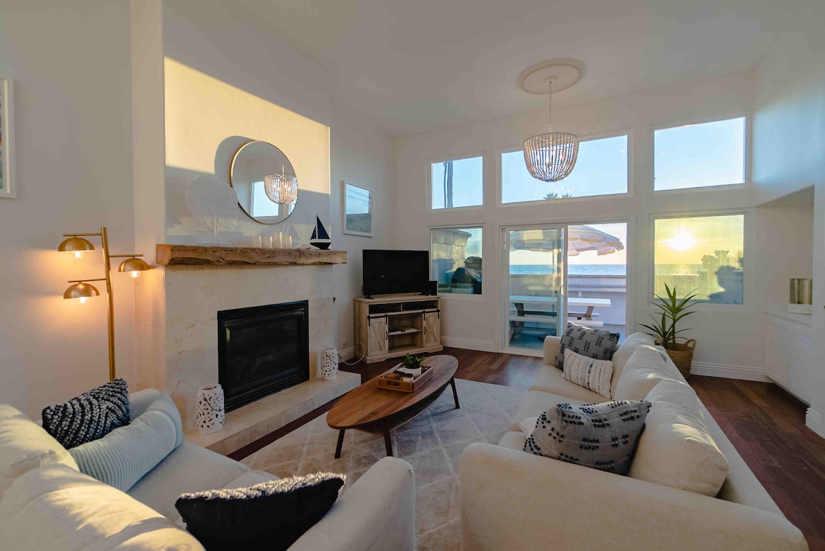 A spacious living area is highlighted by a gas fireplace beneath a large mirror. Soft sofas are arranged for comfort, and large windows offer unobstructed views of the ocean and sunset. Natural light fills the room, enhancing the relaxed atmosphere.