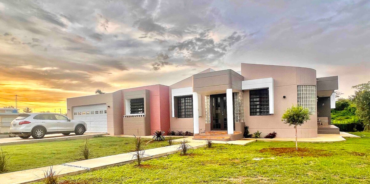 The exterior of a spacious house is displayed, featuring a modern design with a mix of soft pink and beige hues. A neatly landscaped yard surrounds the home, with a driveway leading up to a garage. A vehicle is parked in the driveway, and a small tree adds greenery to the scene.