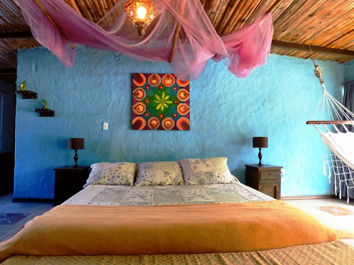 A spacious sleeping area features a large bed with multiple pillows and cozy blankets. Colorful artwork adorns the blue walls, and a hammock is suspended nearby. Decorative elements include lamps on either side of the bed and soft drapery enhancing the ceiling.
