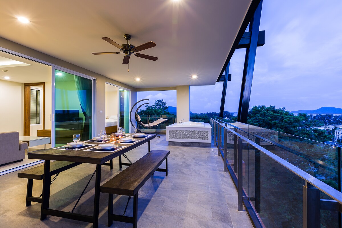 The spacious terrace features a large dining table with ample seating and a luxurious jacuzzi. A contemporary seating area with a hanging chair is visible, alongside sleek glass railings that enhance the view of the surroundings. Soft lighting creates an inviting ambiance.