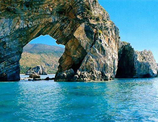 A natural rock formation rising from clear blue waters features a prominent arch. The sun illuminates the rugged texture of the rocks, while distant hills create a serene backdrop. Tranquil waves lap gently at the base of the formation.