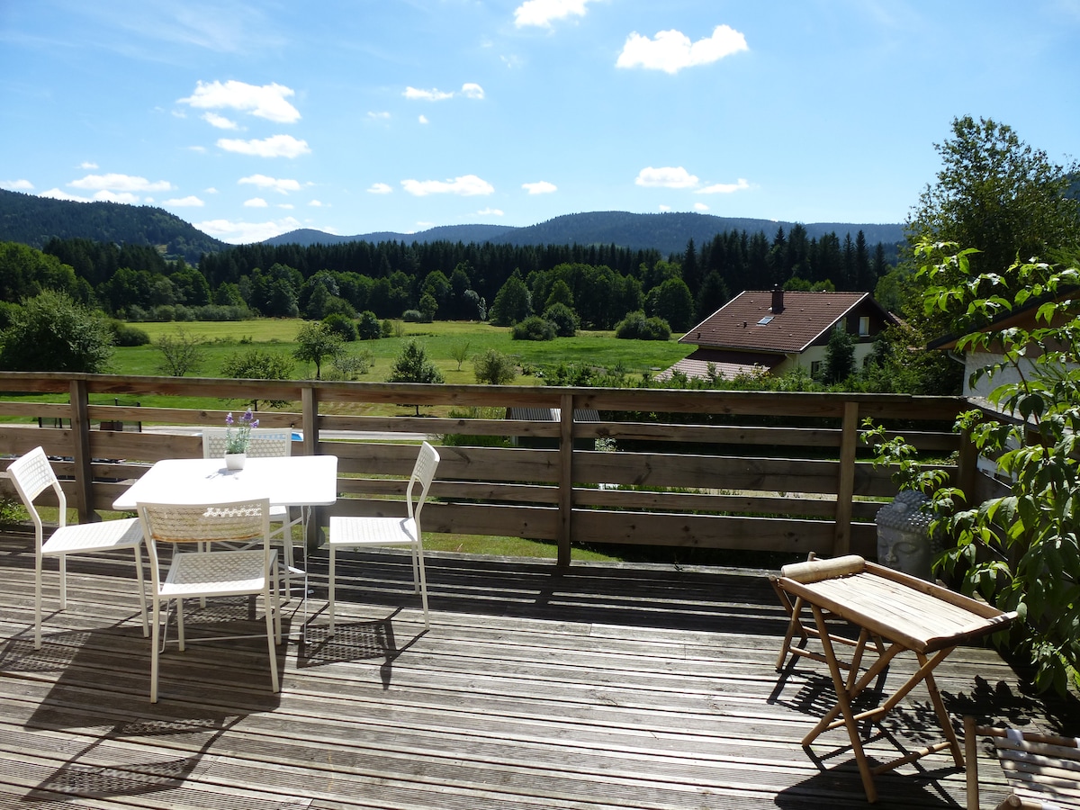 A spacious wooden terrace is presented, furnished with a white dining table and four matching chairs. A folding chair is positioned nearby. Lush greenery and rolling hills are visible in the background, under a bright blue sky dotted with clouds.