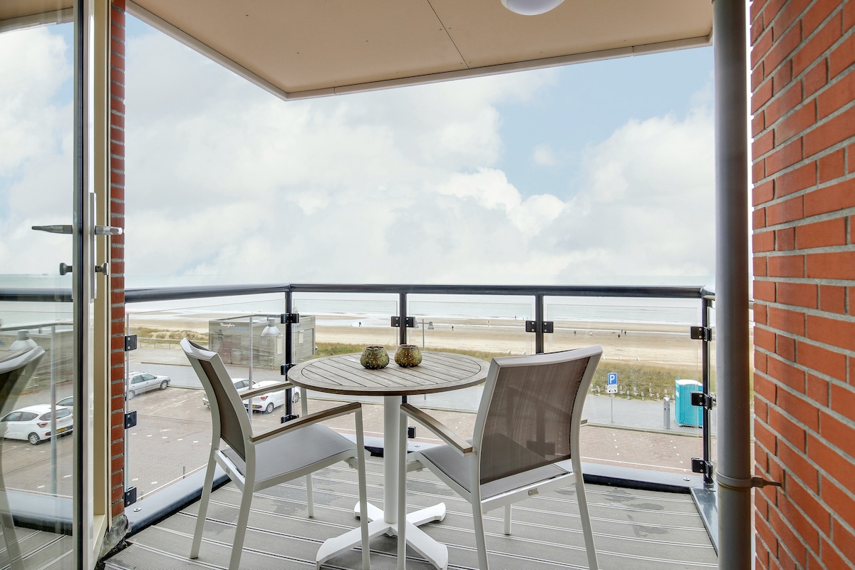 A private balcony is featured, showcasing a round table surrounded by two chairs. An unobstructed view of the beach and sea is visible beyond the railing, inviting relaxation. The space is bright, with soft light illuminating the coastal scene.
