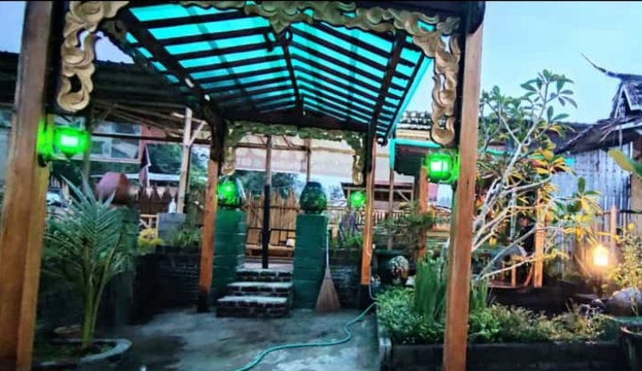 An outdoor area is adorned with wooden arches and a transparent roof, providing a sheltered space. Green lanterns cast a soft glow, illuminating the garden's foliage. Stone steps lead through the greenery, enhancing the serene ambiance of the setting.