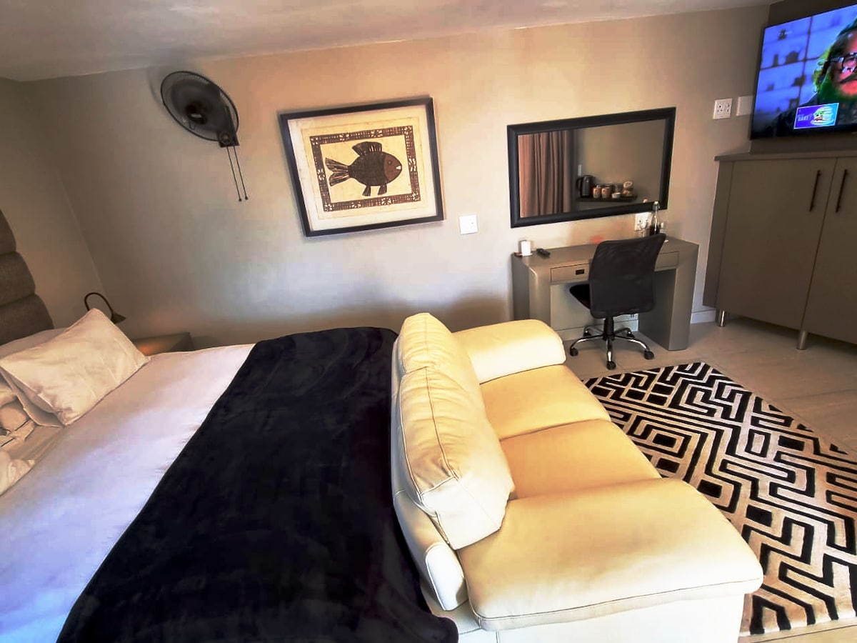 A spacious studio features a king-size bed with a dark throw and decorative pillows. A modern, light-colored sofa is positioned nearby, offering comfort. A desk with a chair is visible, along with a television mounted on the wall, providing entertainment options.