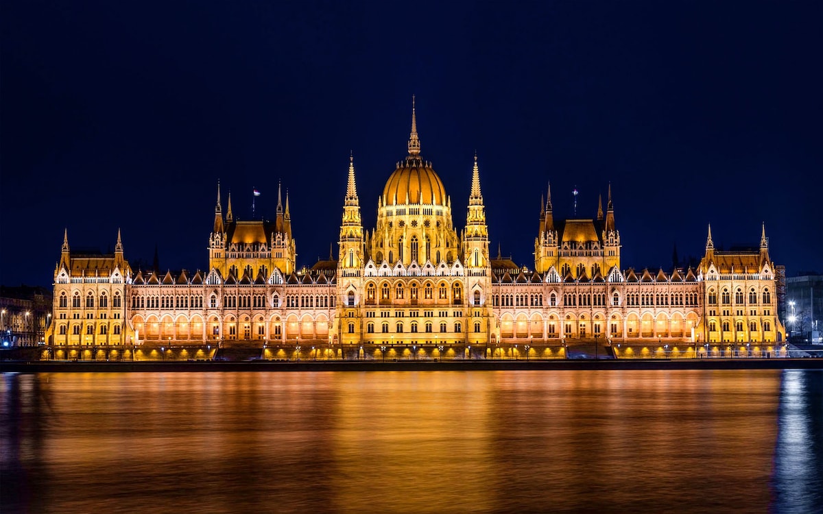 A magnificent historical building is illuminated against the night sky, with its intricate architecture reflected in the calm waters of the Danube River. The structure features domes and spires, showcasing an array of warm golden lights that enhance its grand presence.
