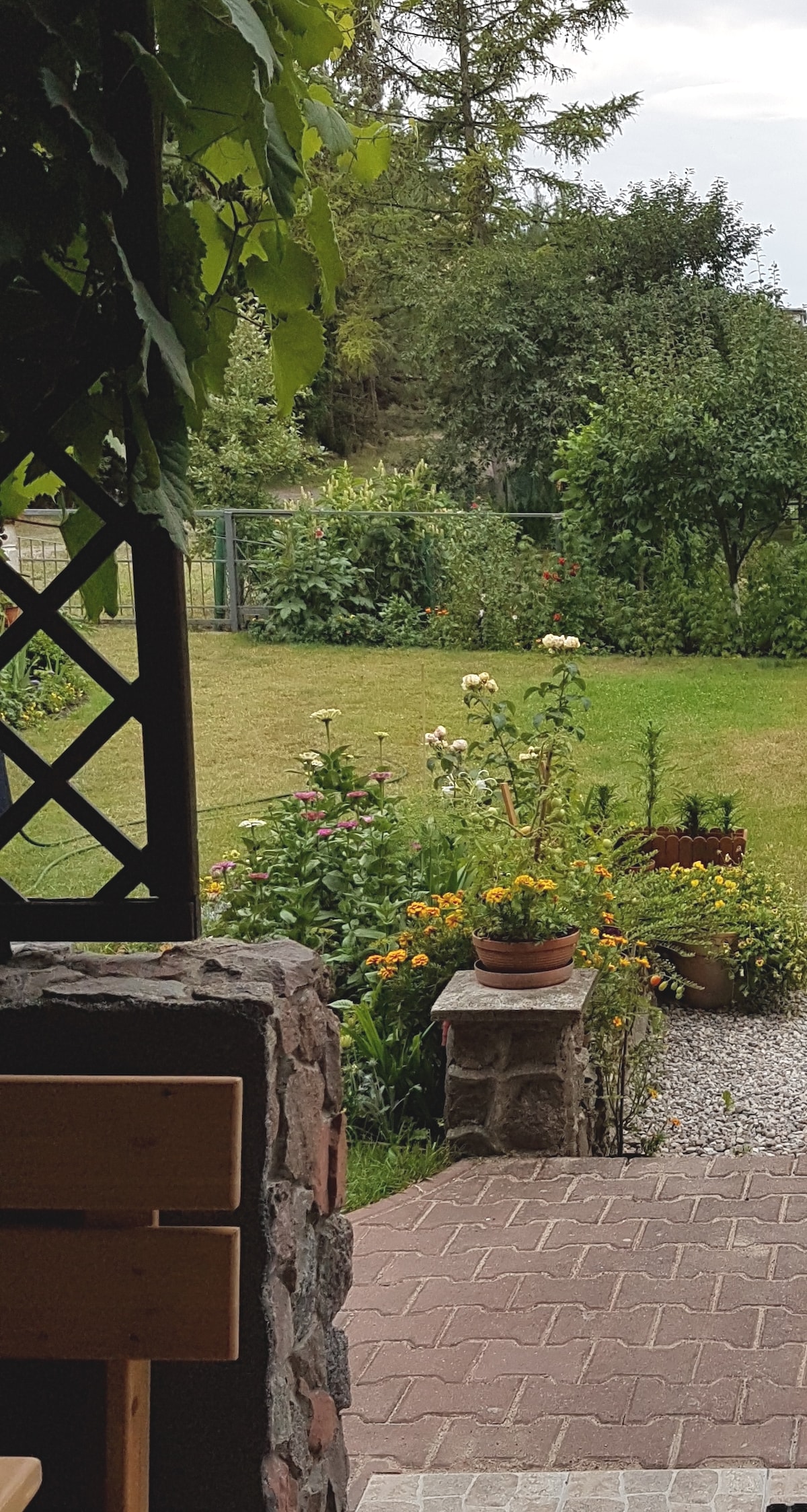 A serene garden view is captured from a covered patio, featuring lush green lawns and vibrant flower beds. Various plants are arranged in pots along the path, contributing to the tranquil setting. The surrounding trees add a sense of privacy and nature.