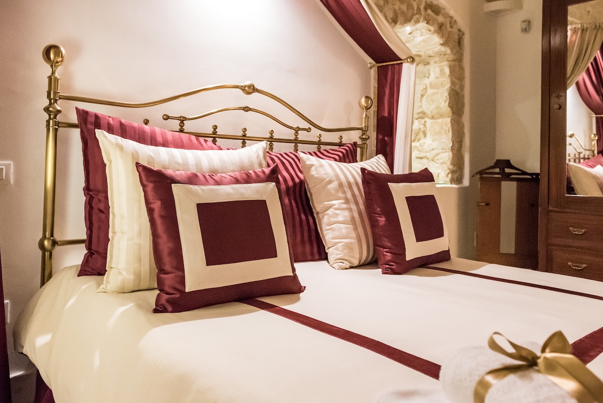 A spacious bed is adorned with decorative pillows in rich burgundy and cream tones. The elegant headboard features a classic design, complemented by subtle textile patterns. Soft, warm lighting enhances the room's cozy feel, while a mirror reflects the carefully arranged furnishings.