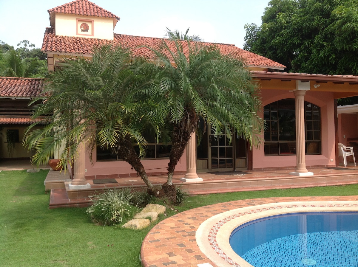 A vibrant exterior view of a spacious house is featured, showcasing a pool surrounded by decorative stone decking. Lush palm trees and manicured grass enhance the outdoor space. The architectural design includes a terracotta-tiled roof and columns at the entrance, creating an inviting atmosphere.