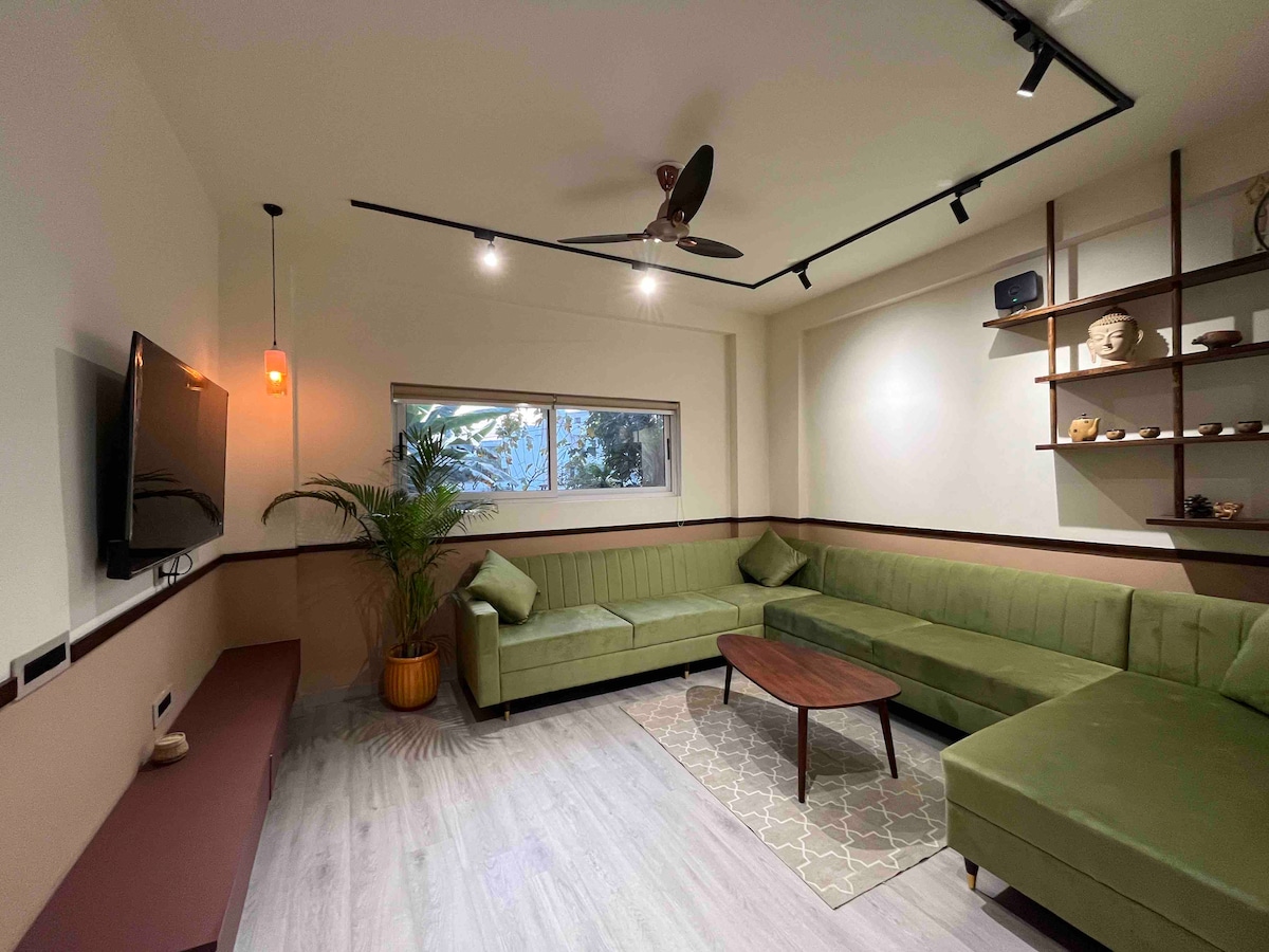 A spacious living area features a green sectional couch arranged around a wooden coffee table. A large wall-mounted TV is positioned opposite the seating. Natural light enters through a wide window behind the couch, and a potted plant adds a touch of greenery to the space.