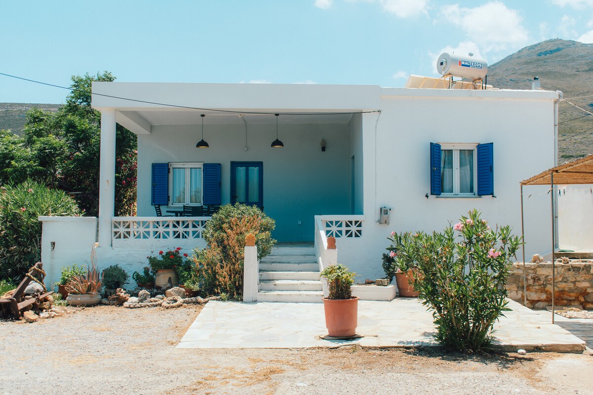 A white single-story house features blue window shutters and a welcoming porch with two hanging lights. Decorative plants are placed along the stairs, and a gravel pathway leads to the entrance. The surrounding landscape offers a glimpse of greenery and distant hills.
