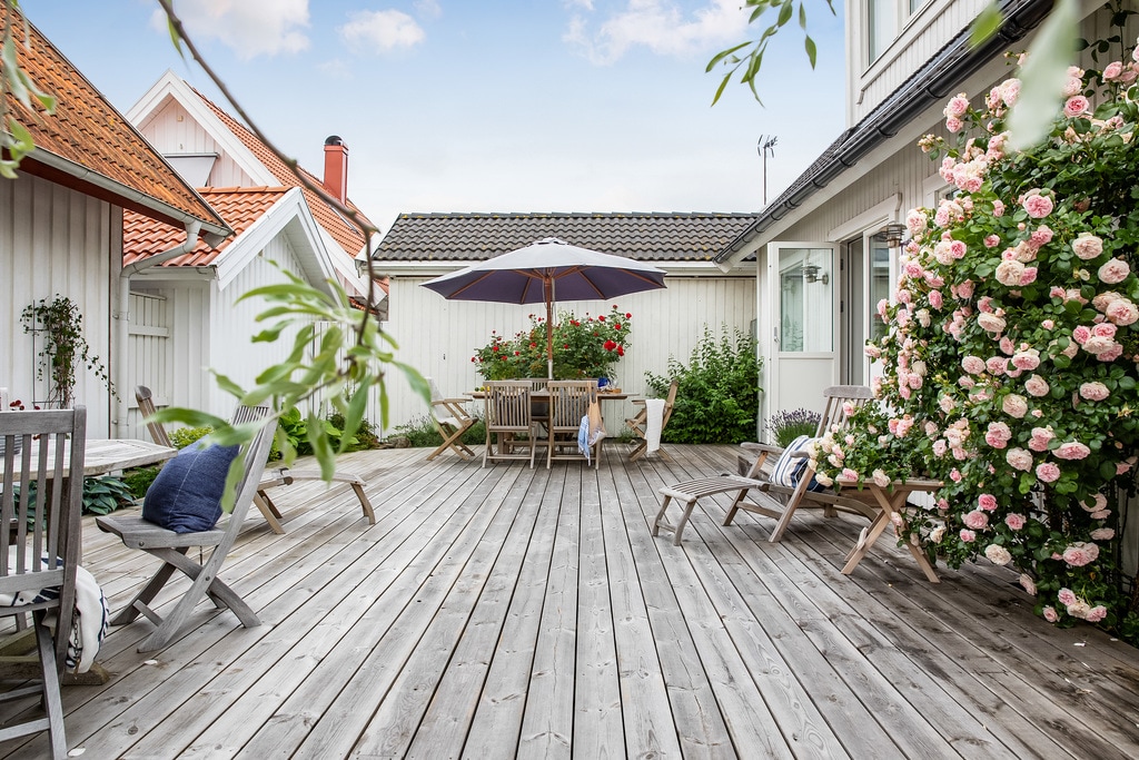 A spacious wooden courtyard is surrounded by flowering shrubs, offering a relaxing outdoor setting. Multiple lounge chairs and a shaded dining table with an umbrella are visible, providing a perfect space for outdoor meals or leisure activities.