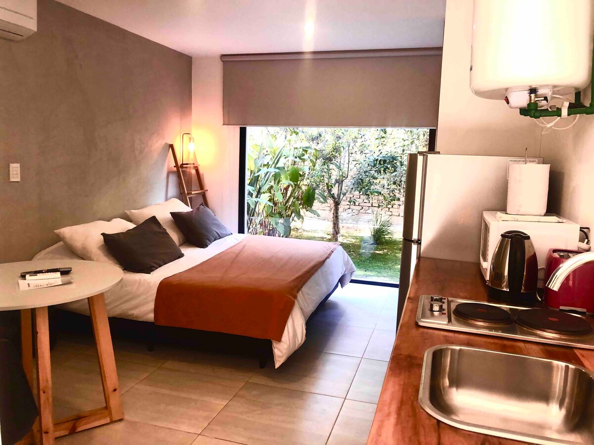 A spacious sleeping area is highlighted by a large bed with neutral bedding and decorative pillows. A small round table with wooden legs is positioned nearby. The space features large glass doors that open to a green outdoor area, allowing natural light to fill the room.