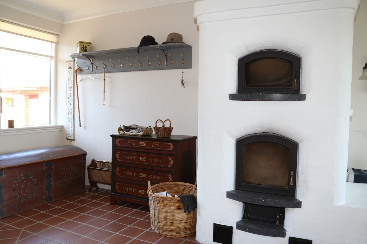 A bright room features a rustic charm with a white brick fireplace and two ovens. A vintage wooden dresser displays intricate designs, while a woven basket and a hat rack with several hooks are positioned nearby. Natural light enters through a large window.