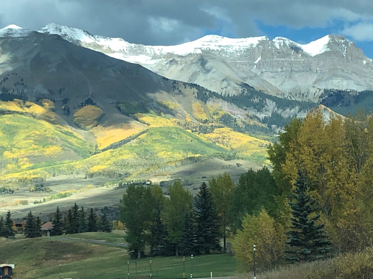 A stunning view showcases majestic mountains with snow-capped peaks in the background. Rolling hills are adorned with vibrant autumn foliage, featuring shades of yellow and green. Evergreen trees are grouped thoughtfully in the foreground, enhancing the natural beauty of the landscape.