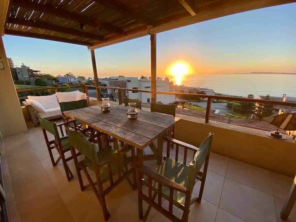 An outdoor terrace is featured, furnished with a wooden table surrounded by six chairs. A warm sunset casts a golden hue across the horizon, while the sea can be seen in the background. Soft seating is arranged for relaxation under a partially shaded roof.