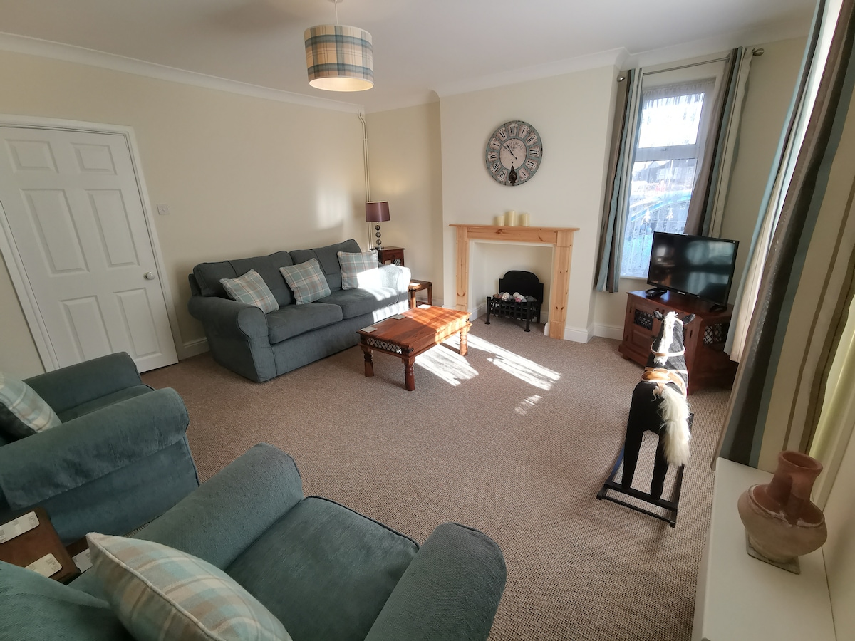 A welcoming living area is filled with natural light, featuring a comfortable sectional sofa and armchairs adorned with soft cushions. A wooden coffee table sits centrally, while a television is positioned beside a decorative fireplace. Warm tones are complemented by light-colored walls and framed windows.