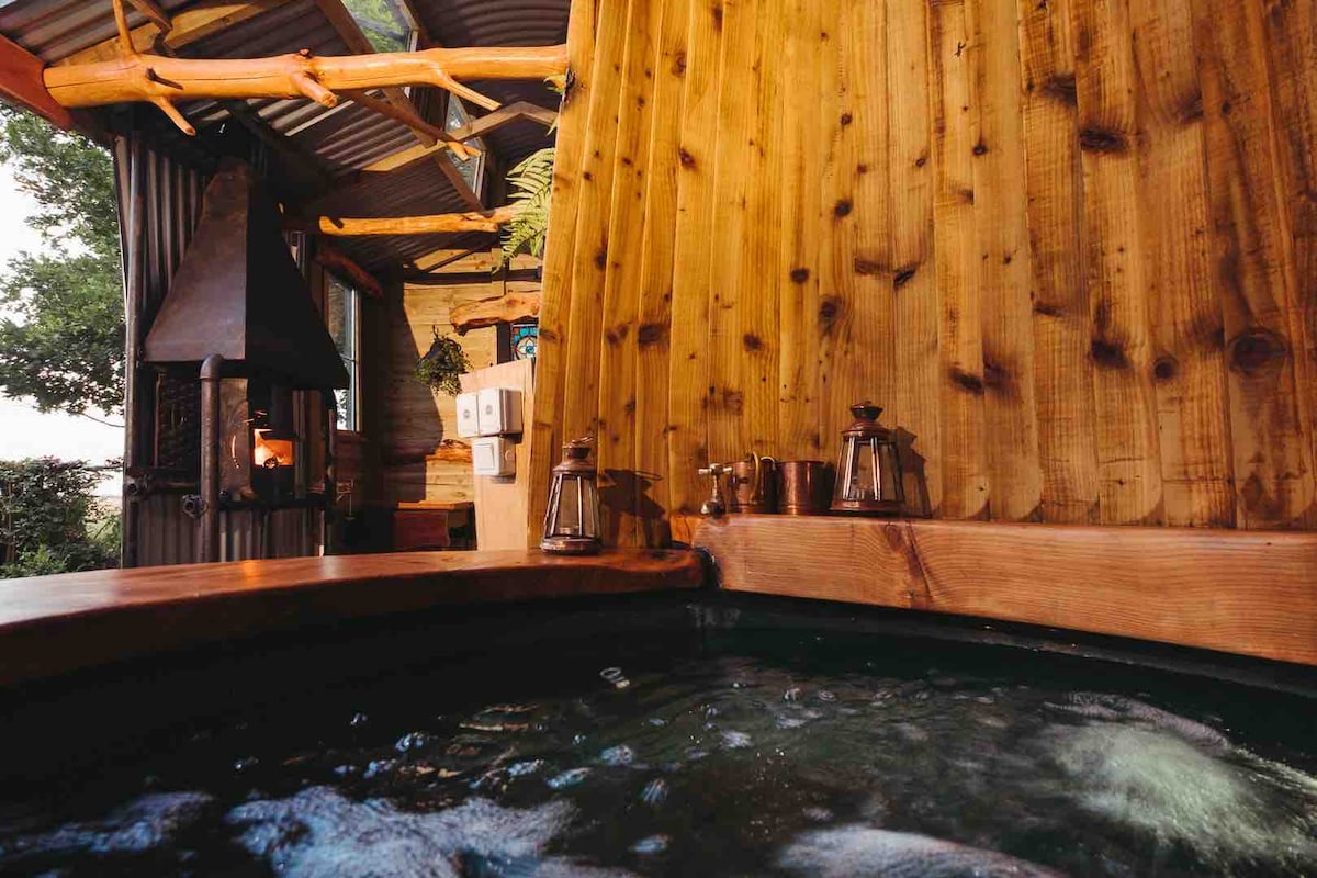 A hot tub made from a large wine barrel is positioned beneath a wooden structure. Warm water is visible, with wood paneling surrounding the tub. decorative lanterns and brass fixtures are placed along the tub's edge, illuminated softly by the ambient lighting, creating a calming atmosphere.