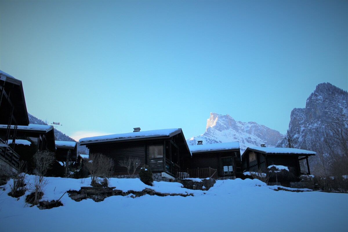 A cluster of charming wooden chalets is nestled within a snow-covered landscape. Majestic mountains rise in the background, under a clear blue sky. The scene conveys a serene winter setting, perfect for a cozy getaway.