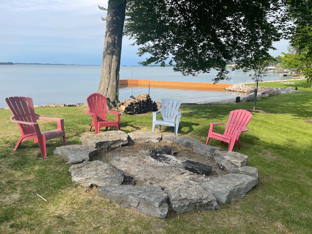 A fire pit area is surrounded by five Adirondack chairs in varying pastel colors, positioned on a grassy lawn. The calm water of the St. Lawrence River is visible in the background, and a large tree offers shade to the gathering space.