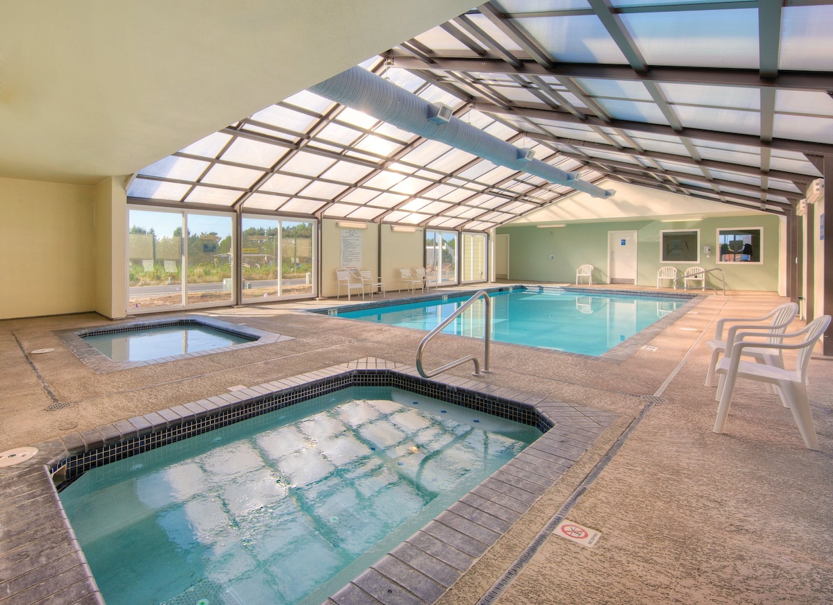 An indoor pool area features a large swimming pool and a hot tub, both illuminated by natural light filtering through the glass roof. White lounge chairs are positioned around the pool, with expansive windows offering views of the surrounding landscape.