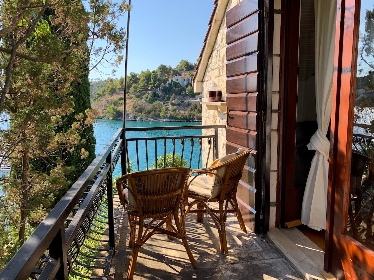 A private balcony overlooks a serene bay, featuring two woven chairs positioned for enjoying the view. Natural stone walls complement the wooden railings, while lush greenery frames the picturesque landscape. Light flows through the open doors into the inviting space.