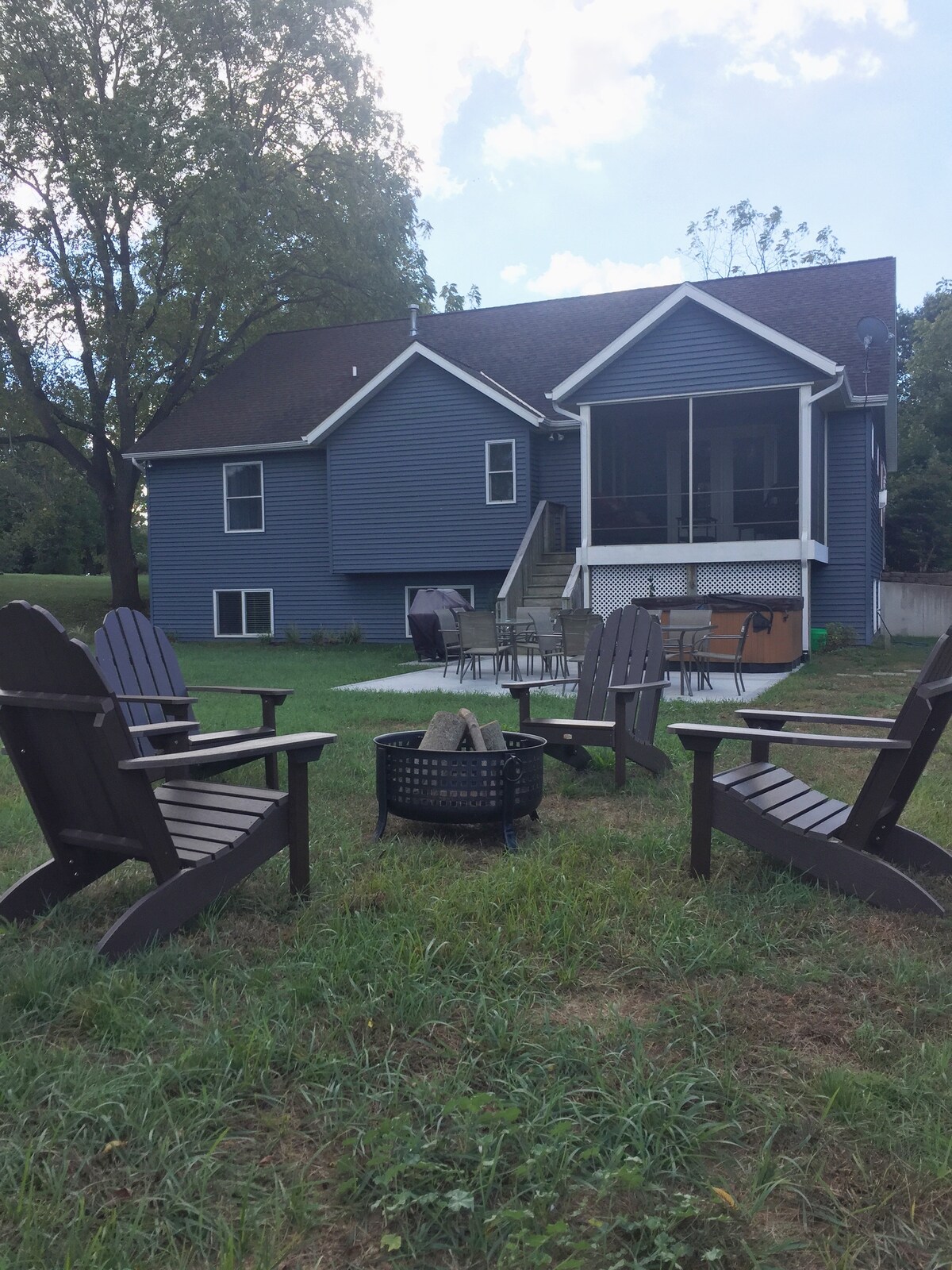 A spacious backyard is highlighted, featuring several brown Adirondack chairs arranged around a fire pit. In the background, a modern blue house is visible, showcasing an elevated screen porch and steps leading to the yard. Lush grass surrounds the seating area.