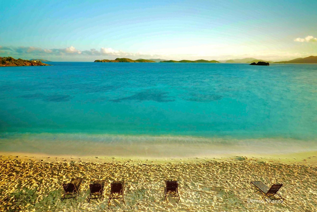 A tranquil sandy beach stretches out, leading to calm turquoise waters. Four lounge chairs are positioned on the shore, offering an ideal spot for relaxation. The distant islands create a serene backdrop beneath a clear sky with soft clouds.