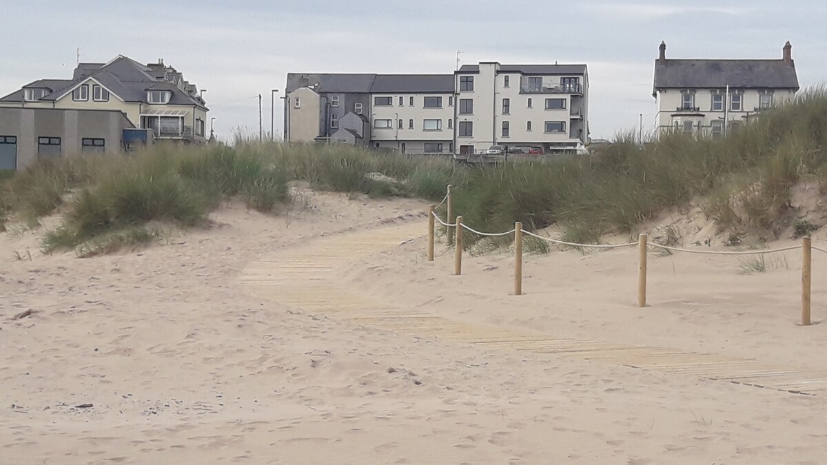 A sandy pathway leads through the dunes towards the beach, framed by tall grasses. In the background, a row of buildings are visible, with a mix of architectural styles. The coastal environment complements the inviting atmosphere of the seaside location.