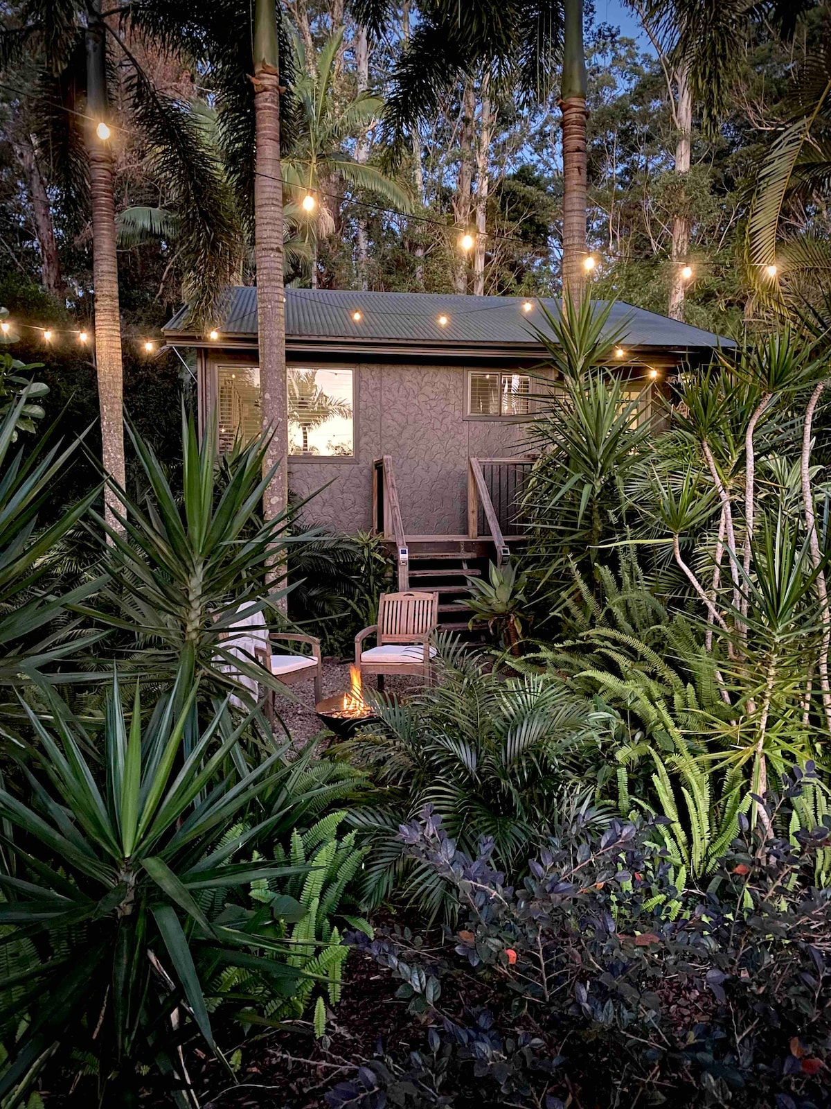 The Aviary cabin is nestled among lush greenery, surrounded by tropical plants. Soft lighting from string lights casts a warm glow, enhancing the inviting exterior. Two wooden chairs are positioned near a fire pit, creating a cozy outdoor seating area within the serene garden setting.