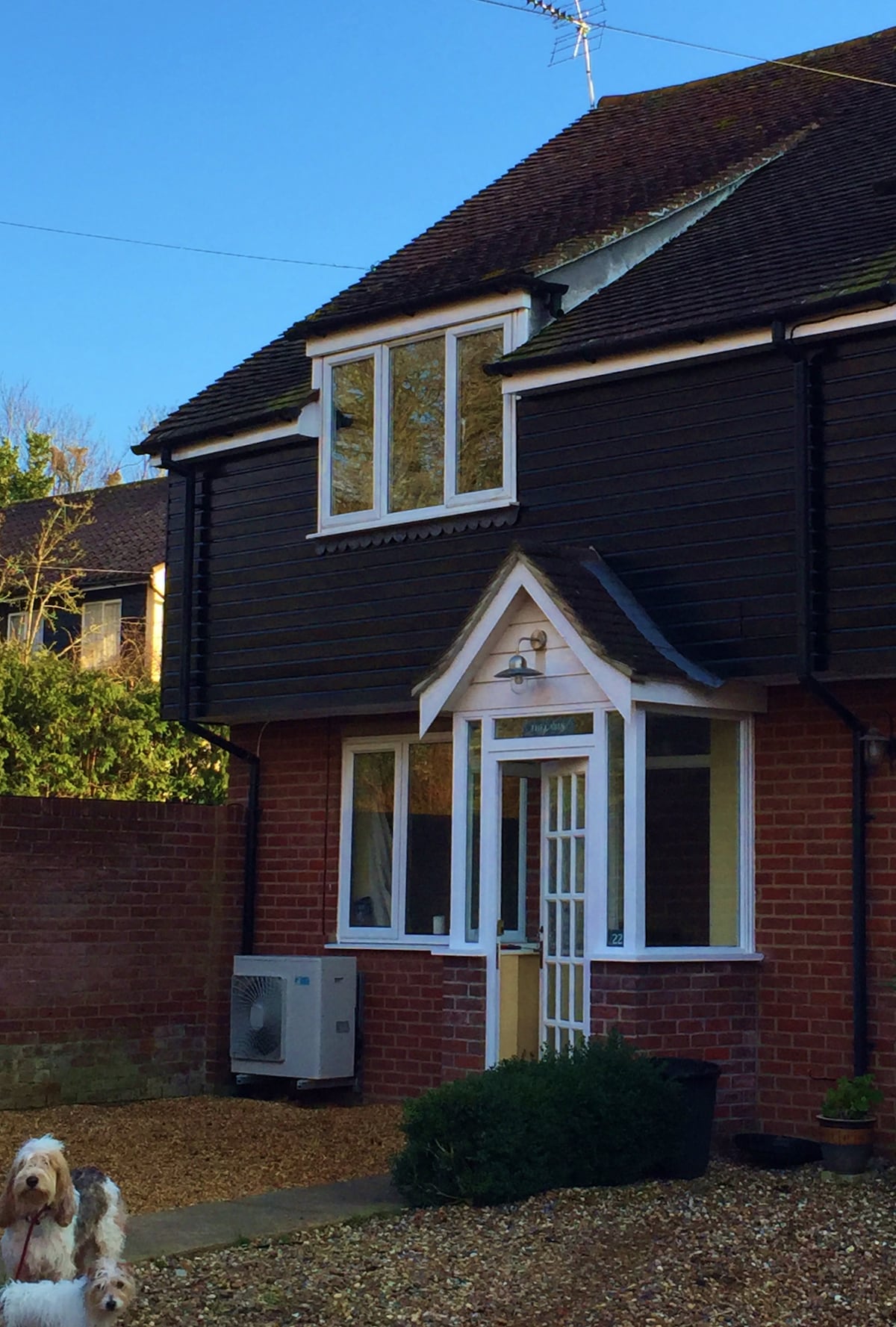 The exterior of the cottage showcases a blend of dark wood and brick, with a welcoming front door framed by large windows. A small garden area is visible, providing a natural touch. A dog is seen in the foreground, adding to the homey atmosphere.