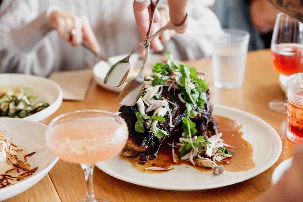 A generous portion of beautifully plated meat is presented on a large white dish, garnished with fresh herbs and vegetables. Clear drinks sit on the table, along with a colorful cocktail in a stemmed glass, enhancing a lively dining atmosphere.