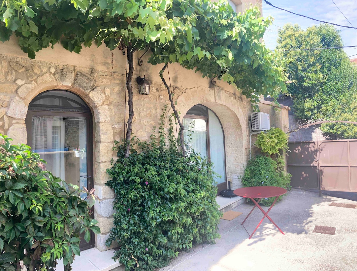 A charming exterior features stone archways adorned with climbing greenery. A small red table is positioned near the entrance, while the ample foliage creates a natural border. Air conditioning units are visible near the entrances, reflecting the warm atmosphere of the setting.
