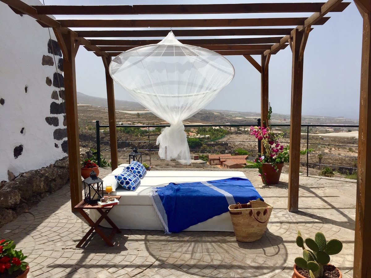 An outdoor terrace features a spacious bed draped with light fabric, providing a serene space for relaxation. Potted plants and a small table adorned with refreshments invite guests to enjoy the stunning surrounding views of the landscape. A woven basket and cacti add a natural touch.