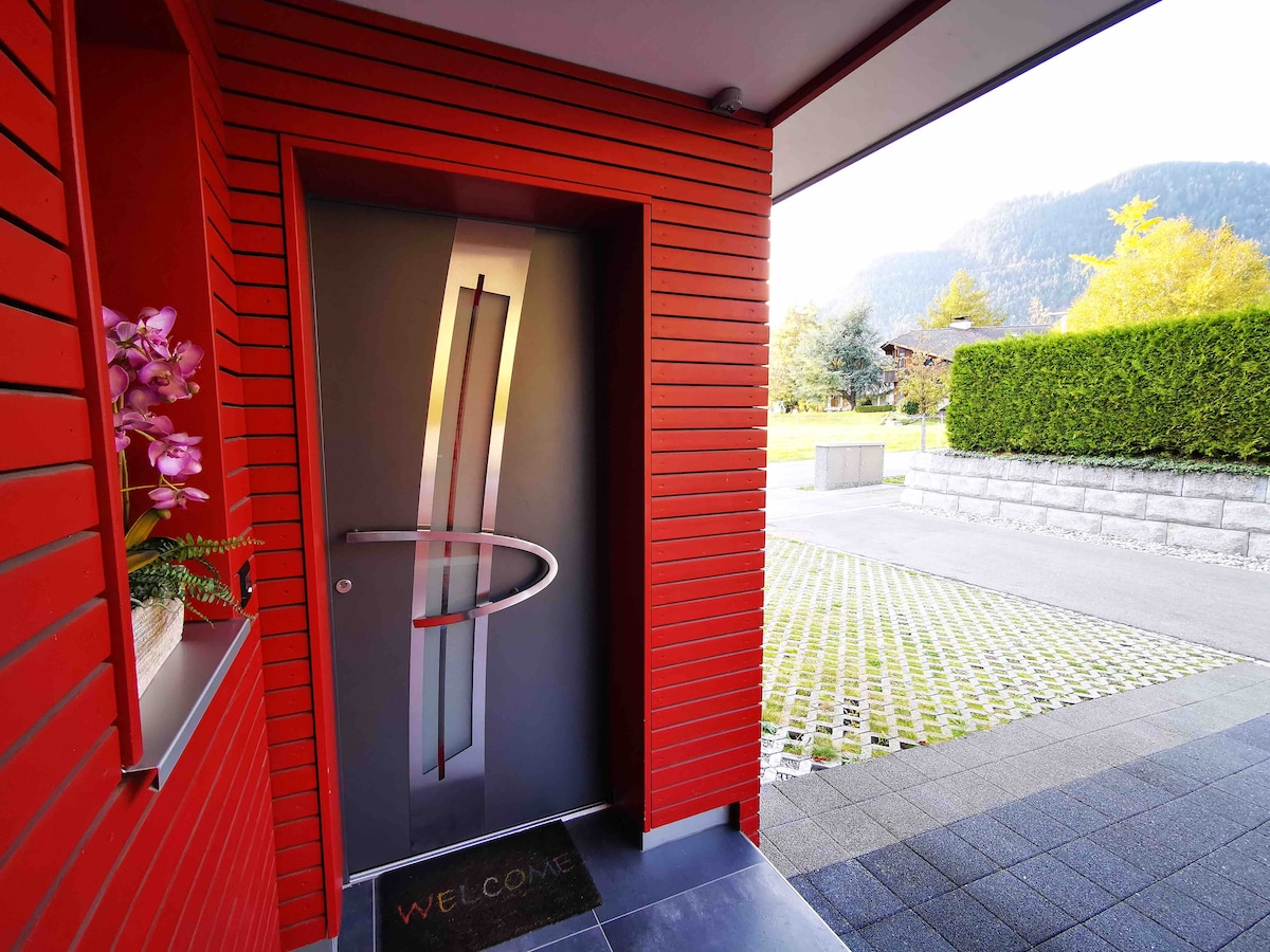 A modern entrance is framed by vibrant red wooden panels. The sleek front door features a contemporary design with a unique handle. A welcome mat is positioned at the base of the door, and a lush garden with green grass and trees is visible in the background.