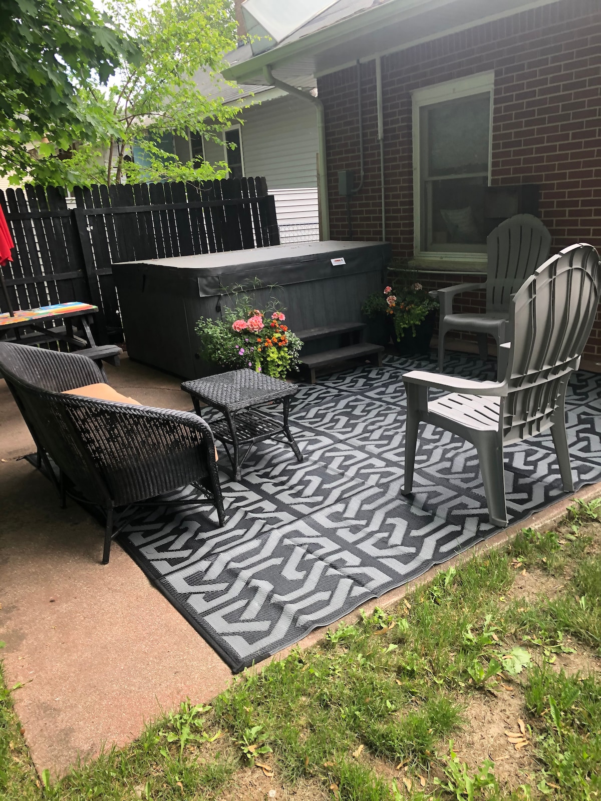A private patio area showcases a hot tub surrounded by potted flowers. Seating consists of two comfortable chairs and a small table, arranged on a patterned outdoor rug. A wooden fence provides privacy while greenery adds a natural touch to the space.
