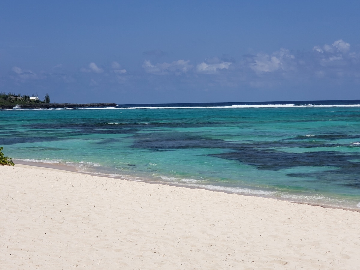 An expansive view of the serene beach is captured, showcasing soft white sand meeting clear turquoise waters. Gentle waves can be observed rolling in the distance, while patches of darker water reveal unique underwater features. A bright blue sky with a few scattered clouds completes the tranquil scene.