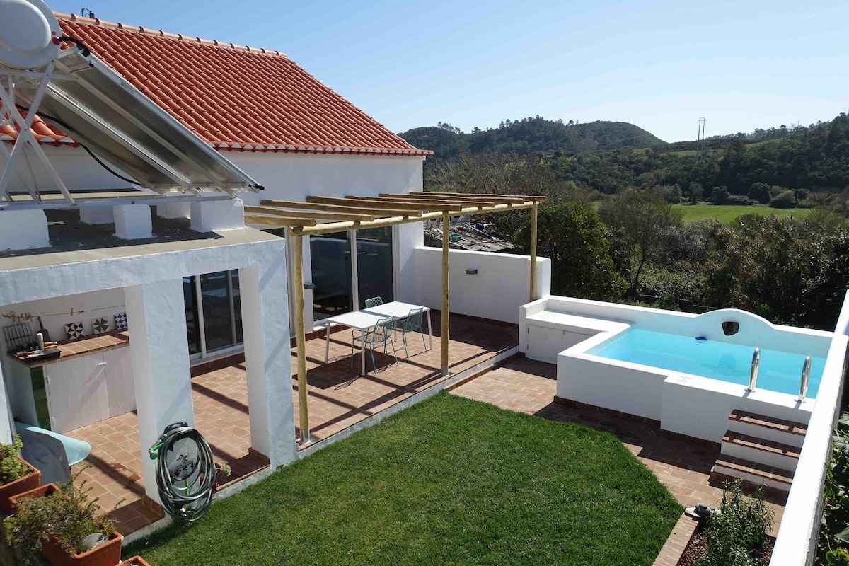 An outdoor area is visible featuring a small swimming pool and a spacious terrace. A wooden pergola provides partial shade over a dining table, while lush green grass surrounds the pool. The white house with a red-tiled roof complements the serene natural landscape in the background.