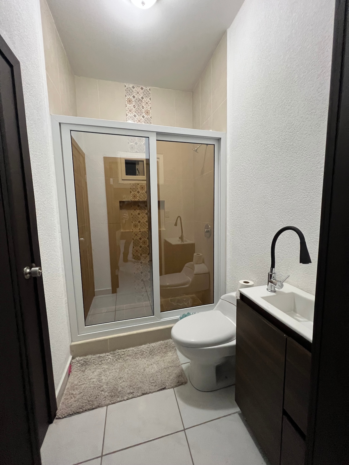 A modern bathroom features a walk-in shower with glass doors, showcasing neutral tiles. A compact vanity with a sink and dark cabinetry is present, accompanied by a plush rug on the tiled floor. Natural light enters through an overhead fixture.