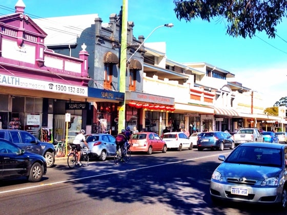 A vibrant street scene is depicted, featuring a mix of shops and cafes in a bustling shopping area. Various vehicles are parked along the road, while cyclists navigate the bike lane, contributing to the lively atmosphere of the community.