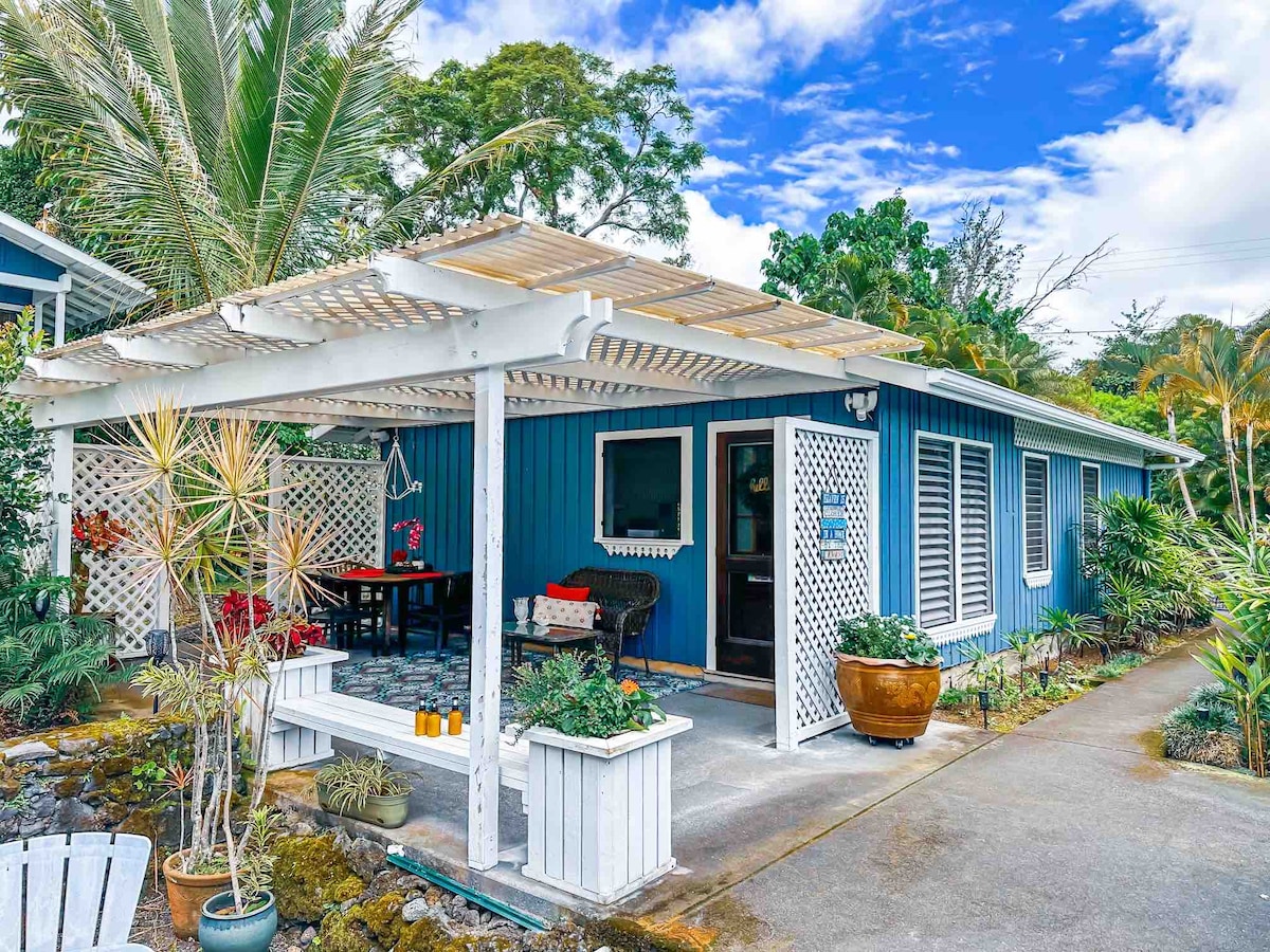 A charming exterior view of the cottage showcases blue wooden siding and a covered porch adorned with tropical plants. A dining area is visible under the pergola, and the surrounding landscaping features various greenery typical of a Hawaiian atmosphere.