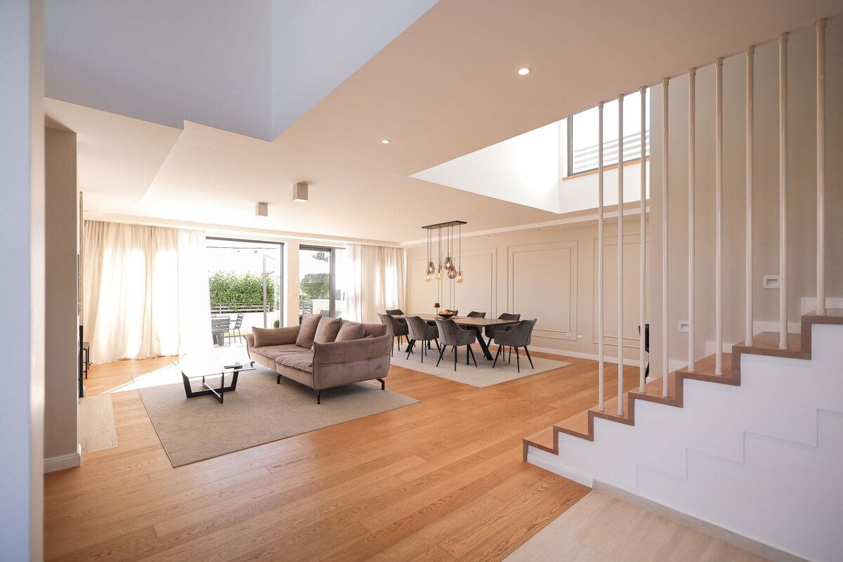 An open-concept living area is highlighted by large windows that allow natural light to fill the space. A comfortable seating arrangement is located in the center, accompanied by a dining area featuring a long table and stylish chairs. Light wood flooring and neutral tones create a harmonious setting.