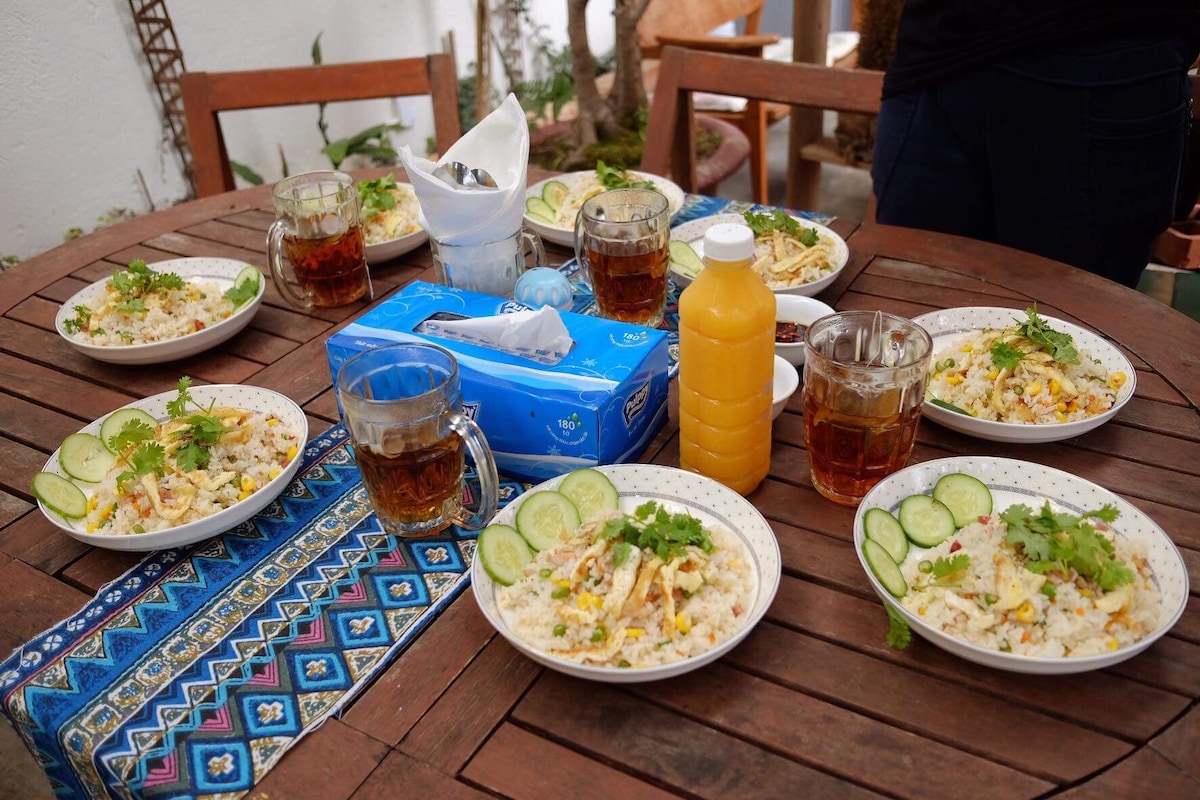 A rustic wooden table is set with multiple plates of rice garnished with fresh vegetables and herbs. Clear glasses filled with a beverage are placed beside each plate. A bottle of orange juice sits in the center, accompanied by a blue tissue box and a folded napkin.