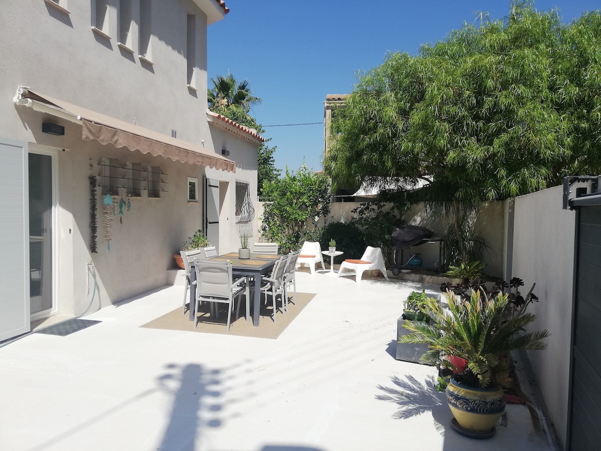 An outdoor terrace features a spacious dining table surrounded by chairs, offering ample seating under the clear blue sky. Two lounge chairs are positioned nearby on the bright, tiled flooring. Lush greenery provides a sense of privacy, enhancing the tranquil outdoor environment.